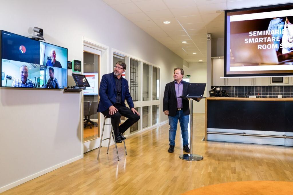 Zwei Männer stehen in einem Raum mit einem großen Bildschirm an der Wand.